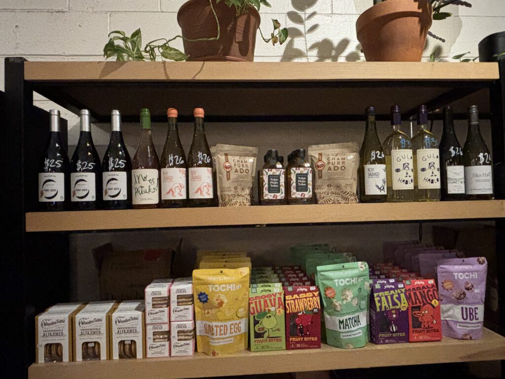 Retail shelf at BookBurn Cafe and Social featuring natural wines, Filipino snacks, and packaged treats displayed beneath potted plants in Orlando.