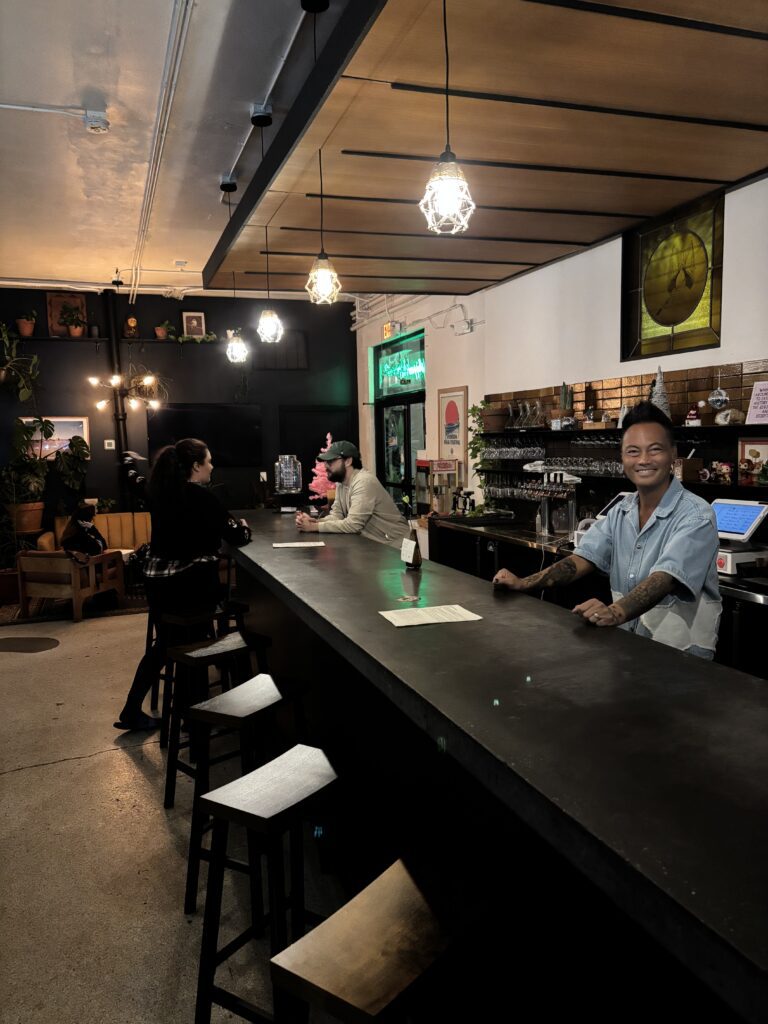 Jonny, the owner of BookBurn Cafe and Social smiling behind the bar at BookBurn Café and Social in Orlando, with guests enjoying drinks in a welcoming community space.