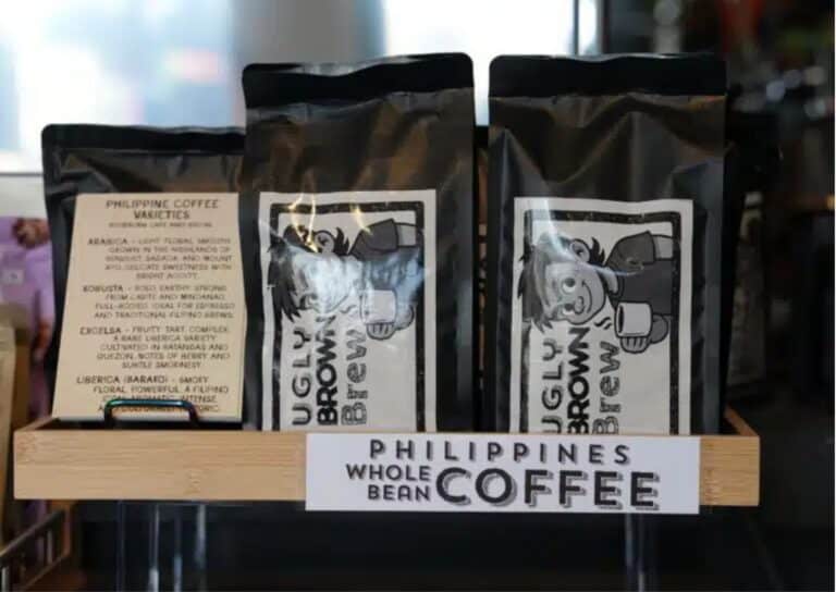Bags of Ugly Brown Brew whole bean coffee on a shelf at BookBurn Cafe and Social, next to a printed card describing Philippine coffee varieties and a "Philippines Whole Bean Coffee" shelf sign.