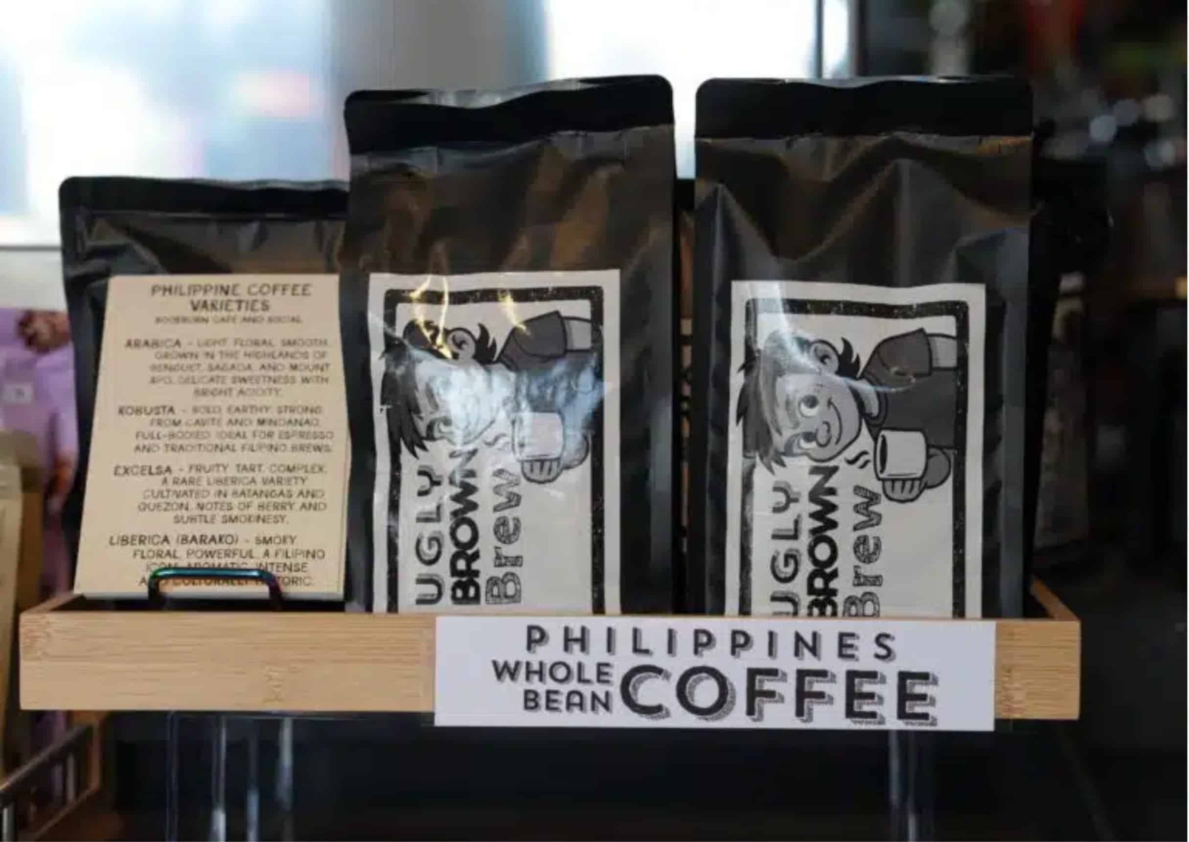 Bags of Ugly Brown Brew whole bean coffee on a shelf at BookBurn Cafe and Social, next to a printed card describing Philippine coffee varieties and a "Philippines Whole Bean Coffee" shelf sign.